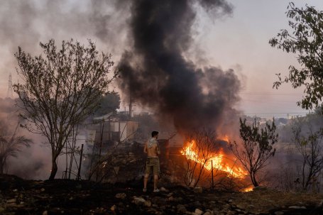 A man in a burned-out area looking at a home on fire.