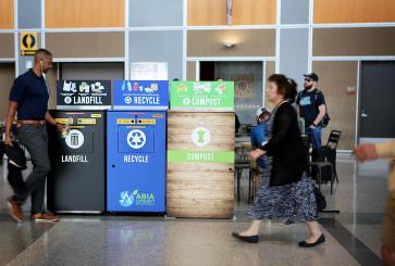 People walk past trash, recycling and compost bins in an airport.