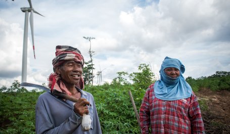 Two farmers stand in their field in Thailand, in front of wind turbines.