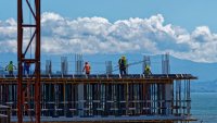 construction workers on top of a building with blue sky, clouds, mountains and the ocean in the background.