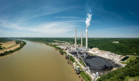 Aerial view of a power plant on a river with piles of coal and smoke rising from smokestacks.