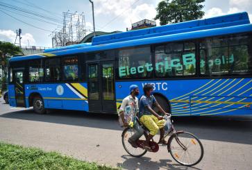 Two people on a bicycle ride past a large blue electric bus in Kolkata, India. 