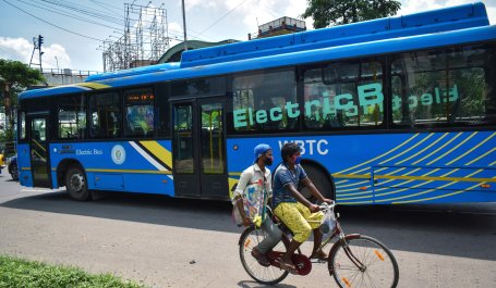 Two people on a bicycle ride past a large blue electric bus in Kolkata, India. 