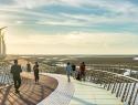 People walk on a bridge over wetlands with windmills in the distance.