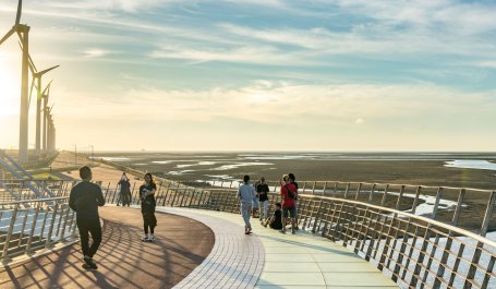 People walk on a bridge over wetlands with windmills in the distance.