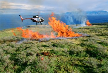 A helicopter flies over burning land in an attempt to extinguish the flames with water.