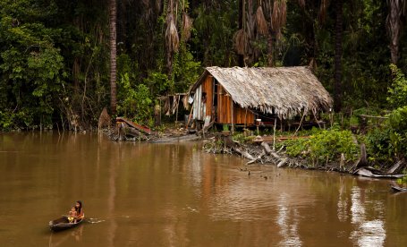 A family in a canoe rows away from a house along a river in the Amazon, Brazil.. 