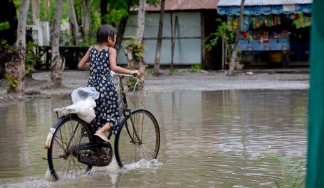 A girl wearing a long sundress pedals a bicycle down a flooded road..