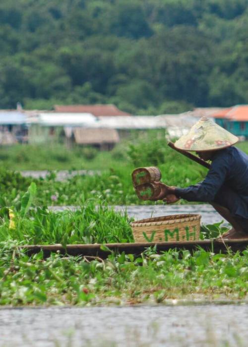 A man fishing on a boat in a river surrounded by fields.