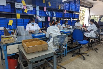 Supply chain workers assemble components while sitting around a long table.