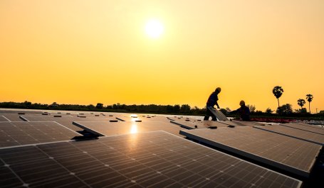 Two workers install solar panels on the corrugated metal roof of a shop.