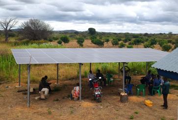 People sitting beneath raised solar panels near a farm field.
