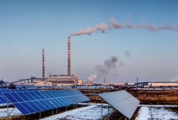 Solar panels with smoke from a smokestack in the background.